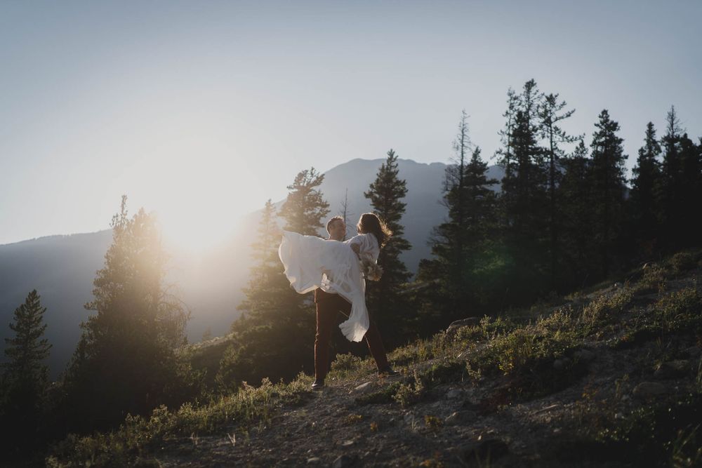 Silhouettes of a couple dancing against a sunlit mountain backdrop with evergreen trees.