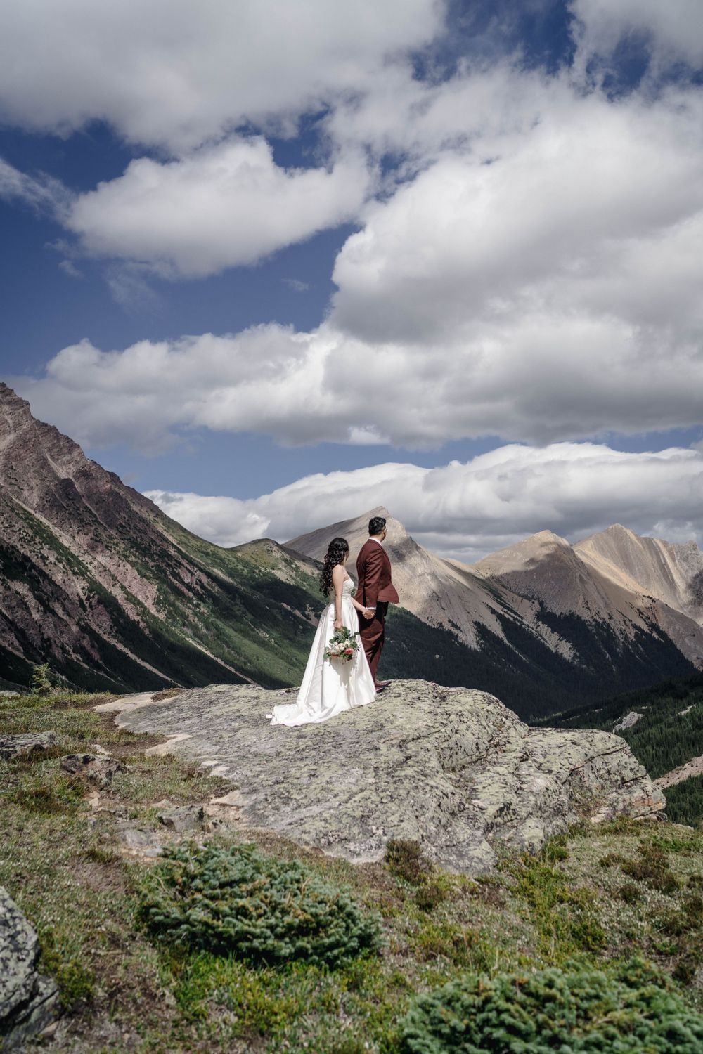 Wedding couple stands on mountain peak overlooking dramatic mountain range under cloudy sky.
