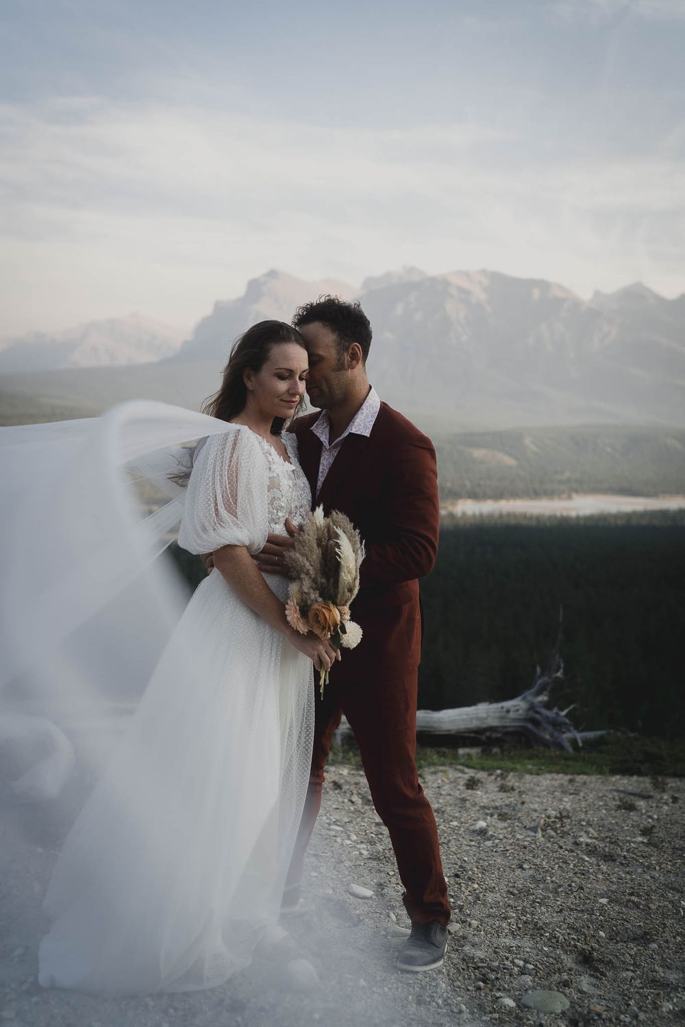 A romantic mountain wedding photo with dramatic views of peaks and forests in the background.