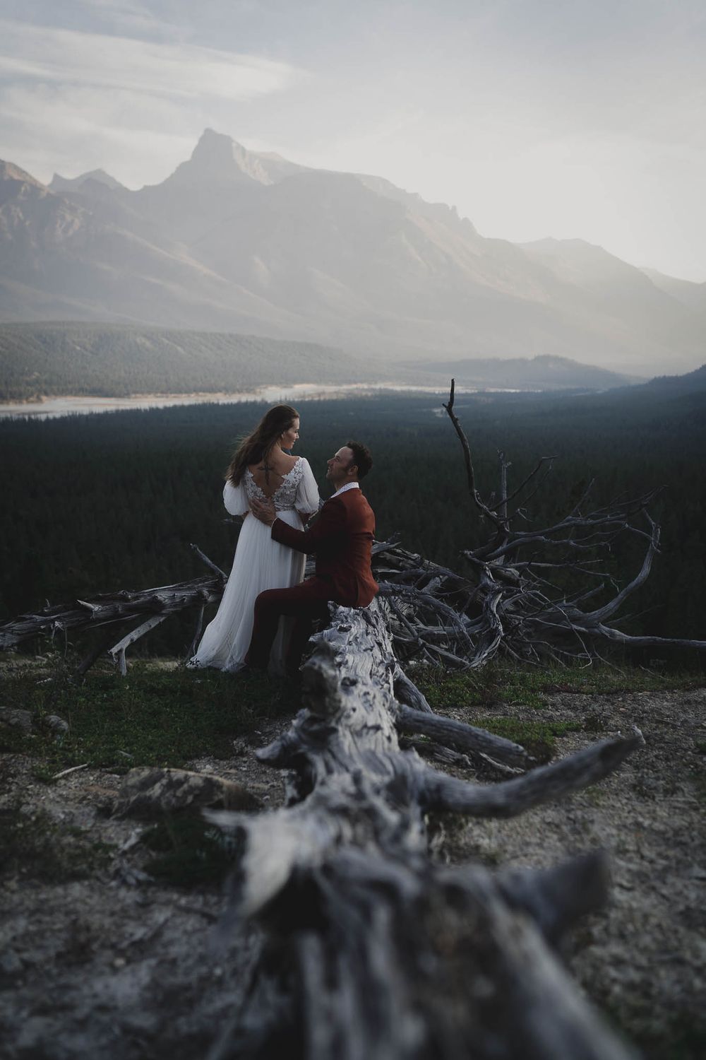 A couple shares an intimate moment on a mountain cliff with fallen trees and scenic valley views.