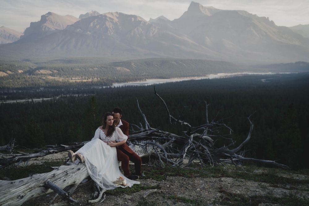 Couple sitting on fallen logs at mountain overlook with dramatic valley and peak views.