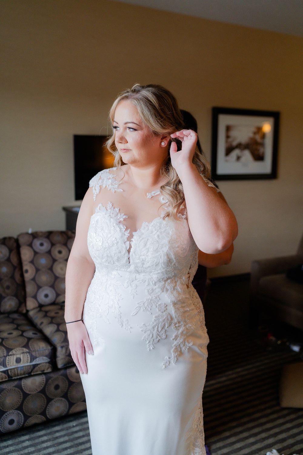 A bride in a fitted lace wedding gown adjusts her earring while getting ready.