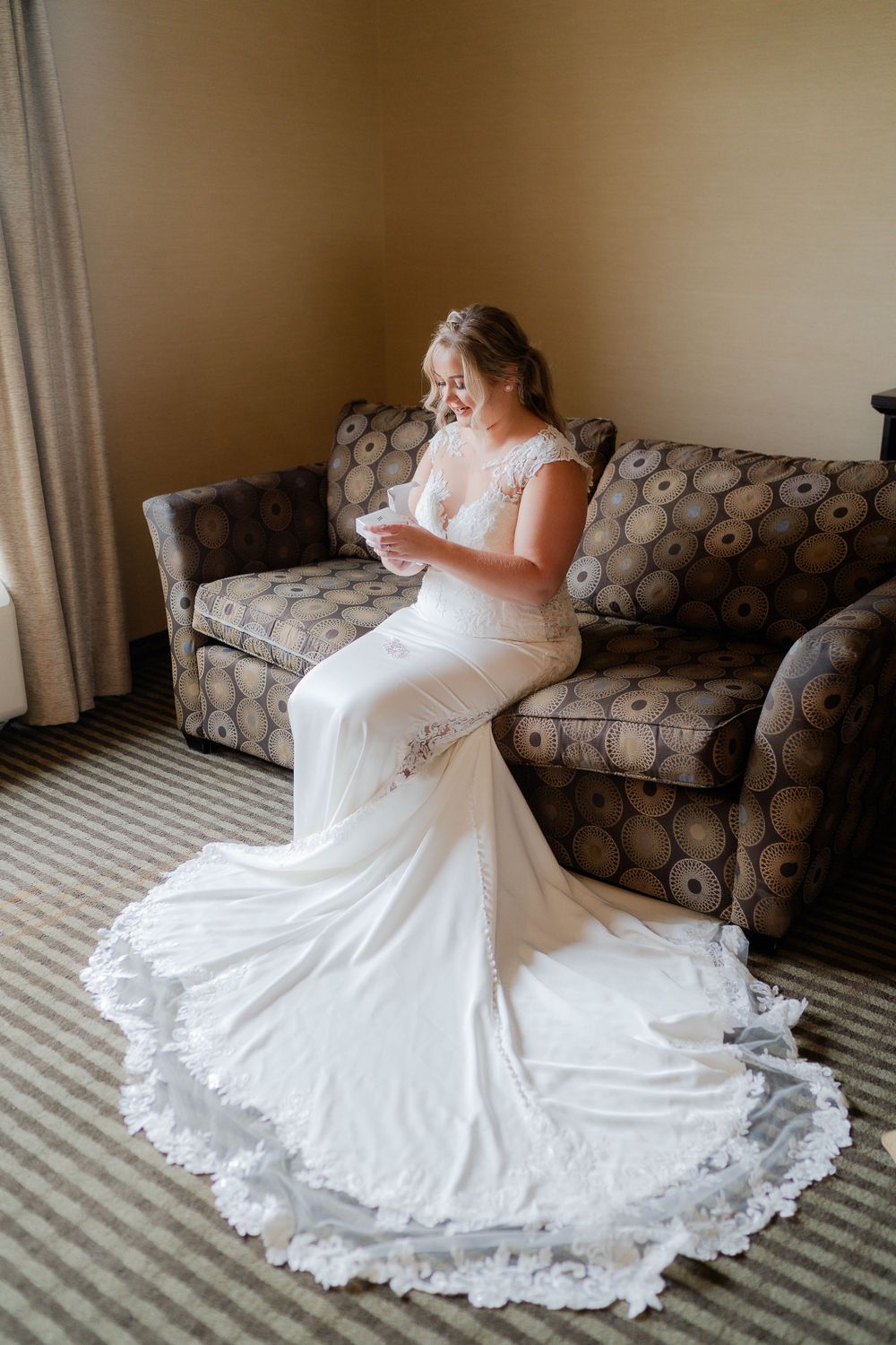 A stunning lace wedding dress with a flowing train displayed elegantly on a patterned couch in natural window light.