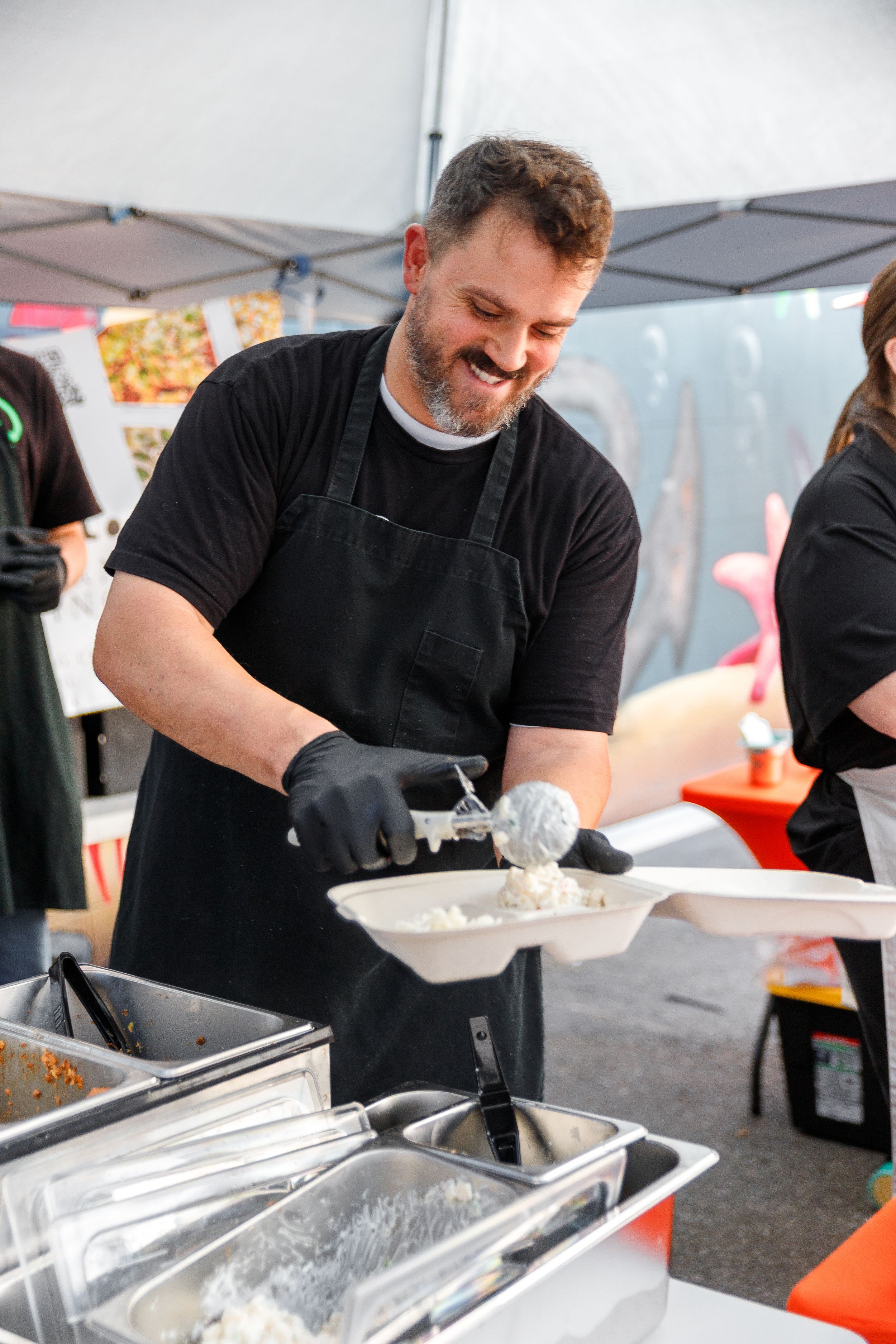 A chef in black apron and gloves serves food at an outdoor event while smiling warmly.