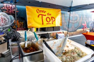 Food vendors serve a variety of dishes at an outdoor market with colorful yellow awnings and signs.