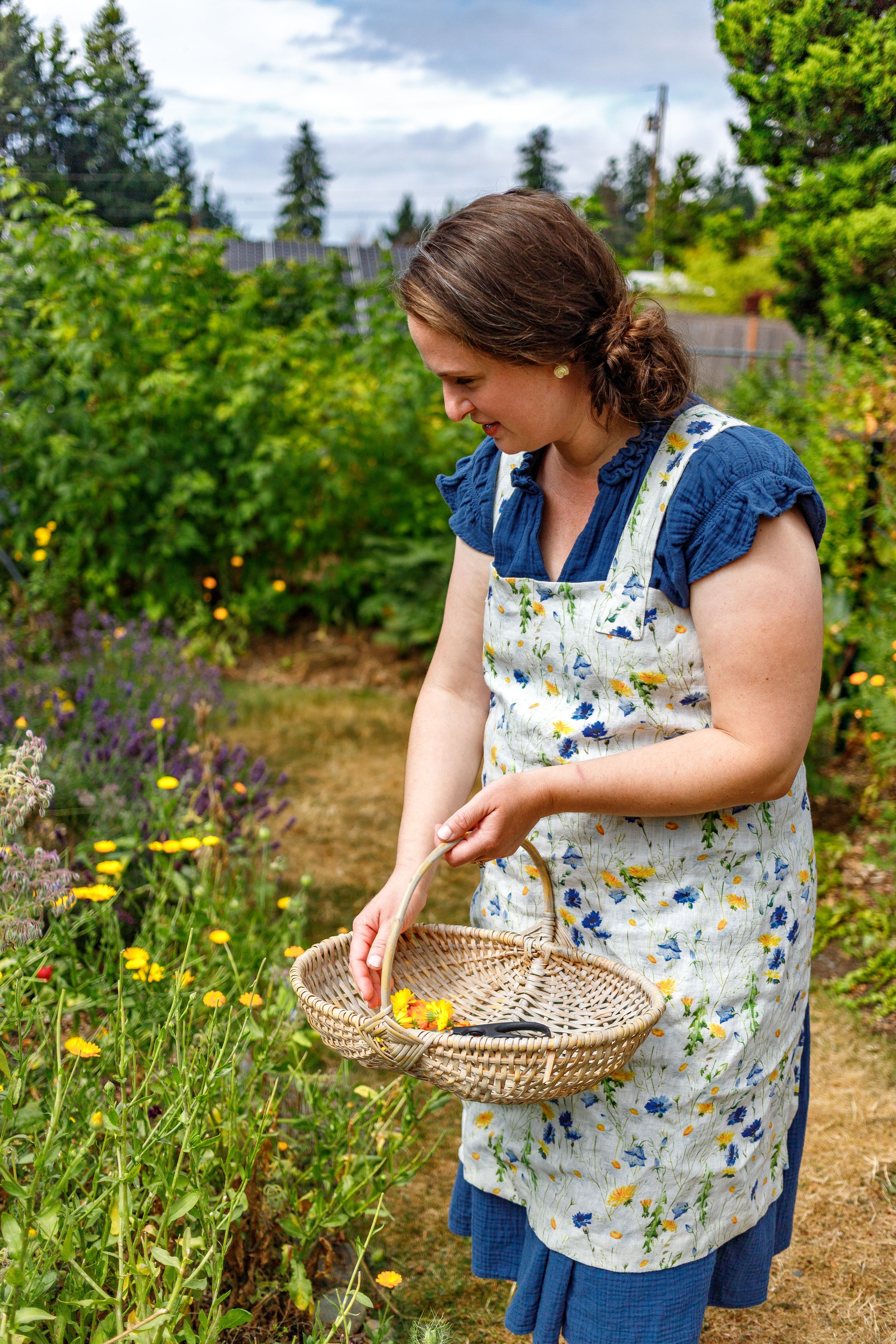 A person wearing a floral apron collects flowers in a woven basket while standing in a wildflower garden.
