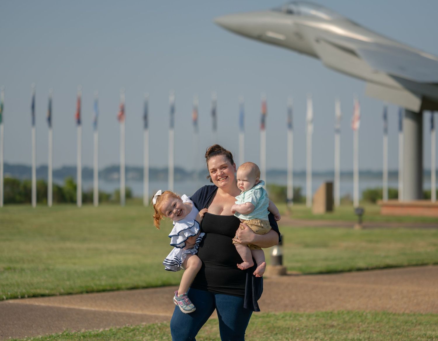 A parent holds two small children outdoors with flags and an aircraft display in the background at a military memorial.
