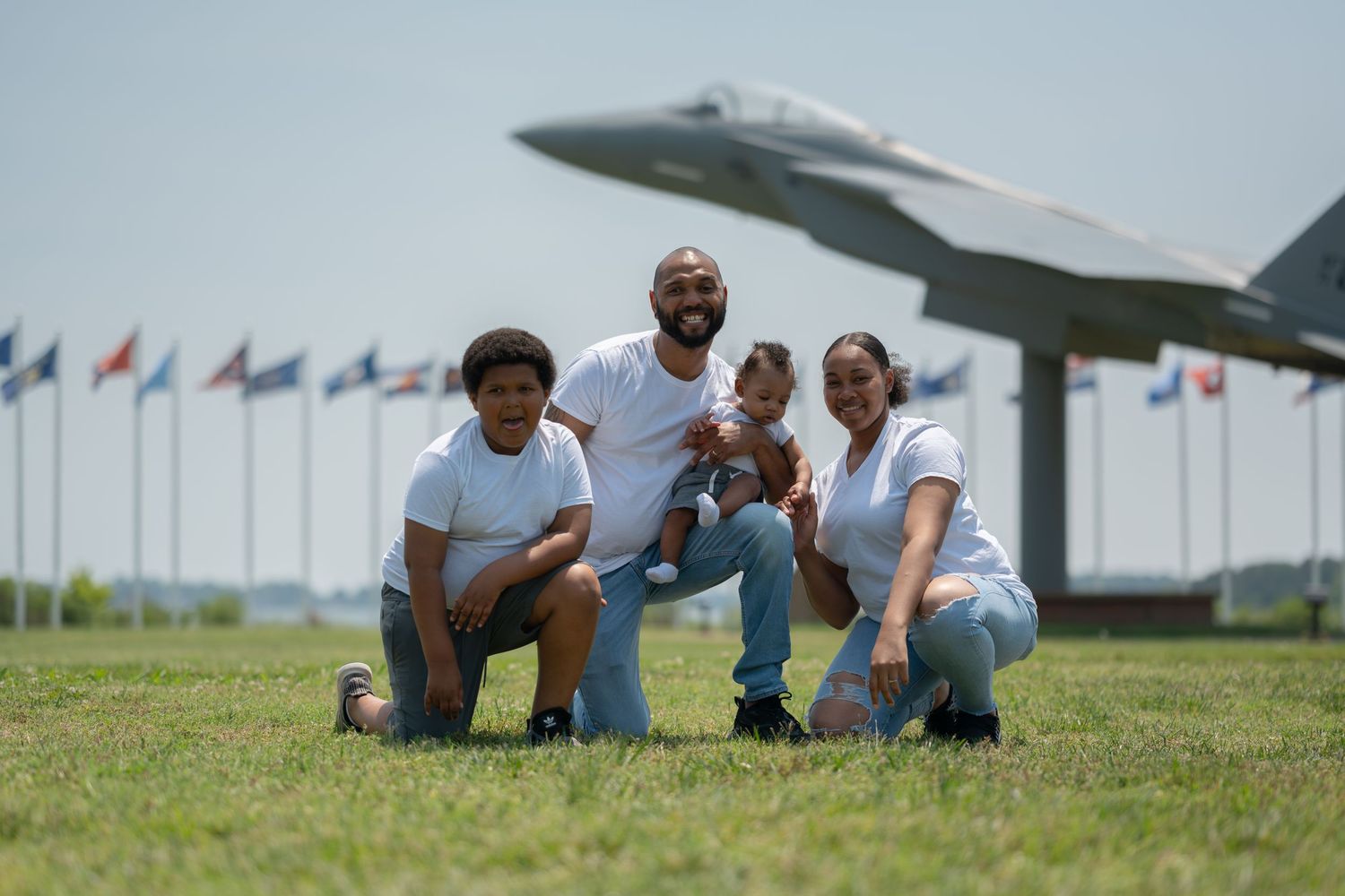 People posing on grass field with military aircraft and flags in background.