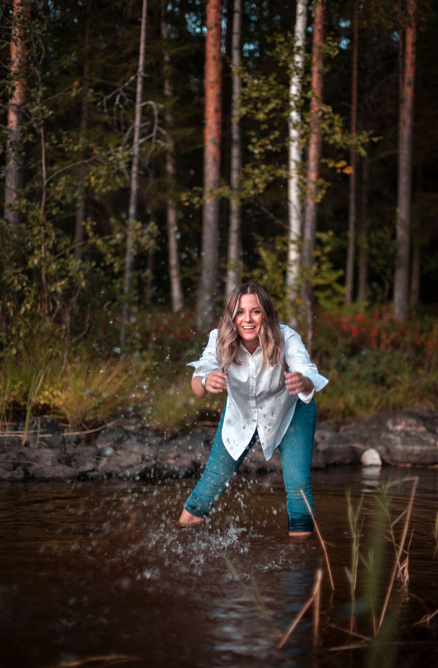 Happy woman splashing in forest stream wearing white shirt and jeans among autumn trees.