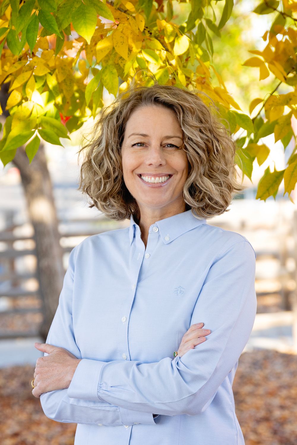 Outdoor autumn portrait with yellow leaves and fence in background, wearing light blue collared shirt.