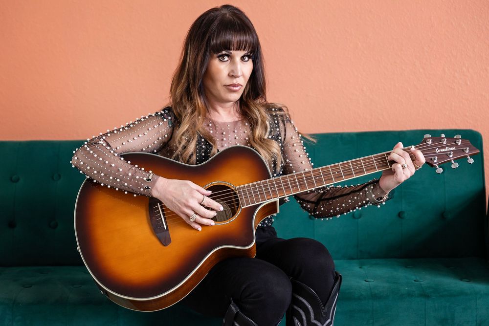 Musician playing sunburst acoustic guitar while sitting on green couch wearing sparkly top.