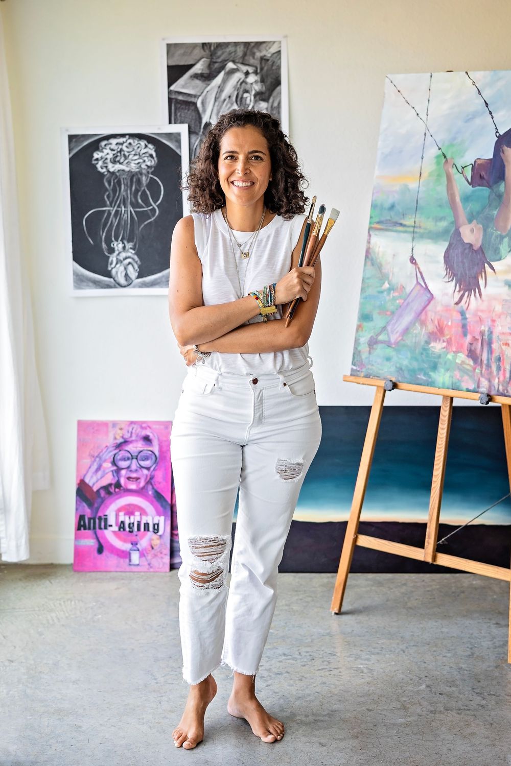 Artist stands barefoot in white attire in studio space with artwork displayed including colorful paintings and sketches.