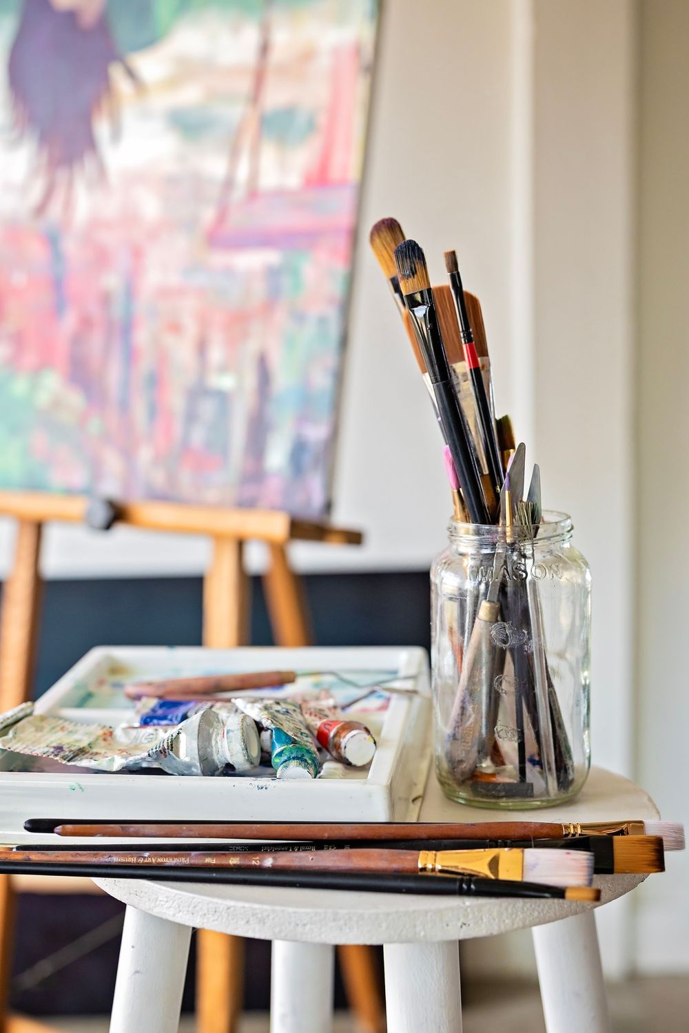 Paint brushes in a glass jar and art supplies on a white stool with colorful artwork in the background.