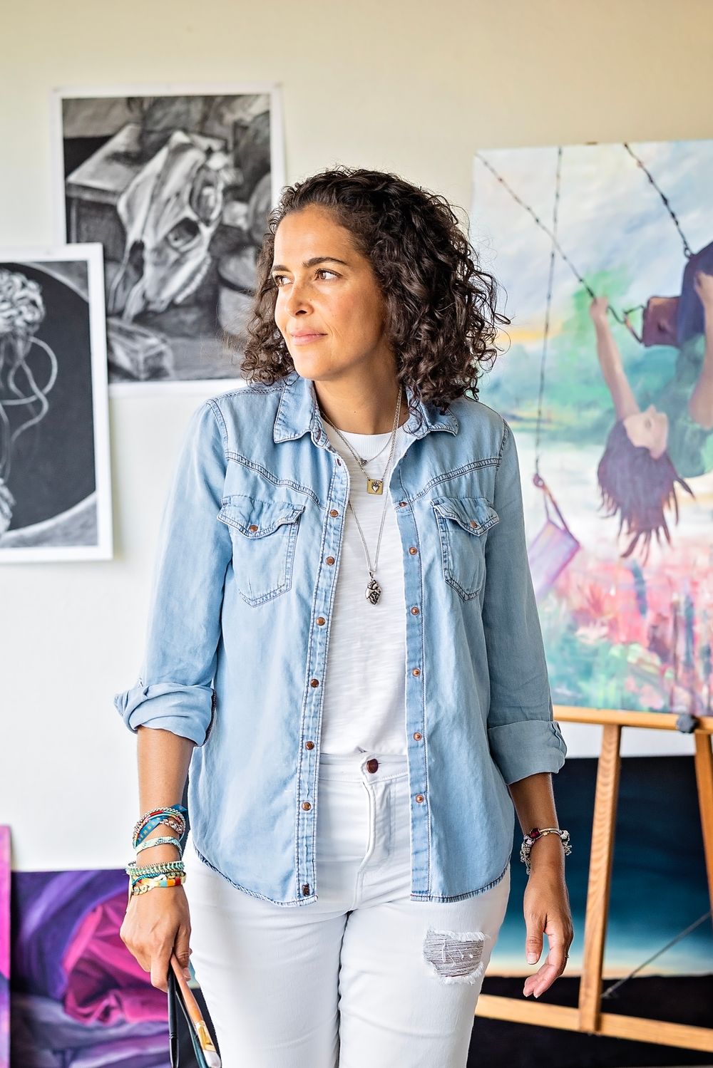 Artist in denim shirt and white jeans stands thoughtfully in studio with colorful artwork displayed on walls.