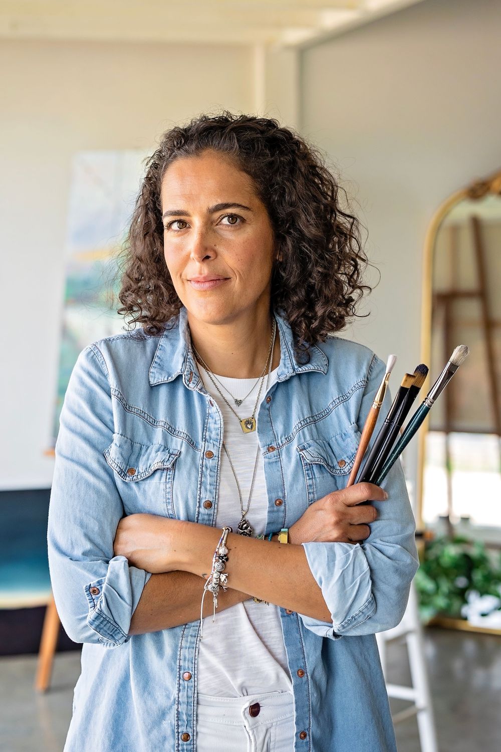 Artist holding paint brushes while standing in an art studio with easels and paintings visible.