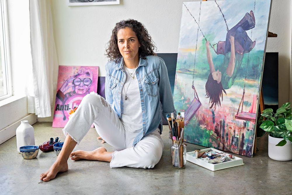 An artist sits on the floor of a studio space with colorful paintings, art supplies, and houseplants visible in the background.