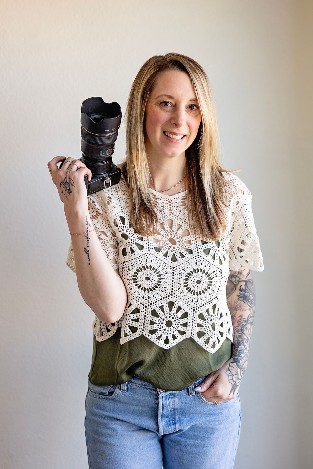 A photographer wearing a white crochet top and jeans holds a professional camera while standing against a neutral wall.