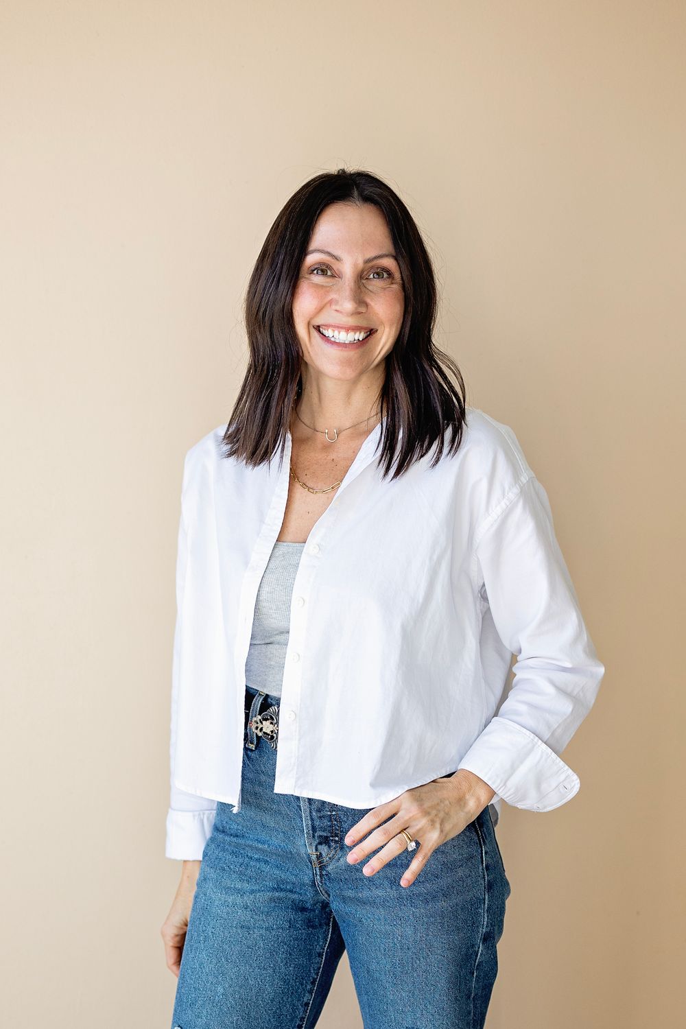 A person wearing a white cropped jacket and blue jeans poses against a beige wall.