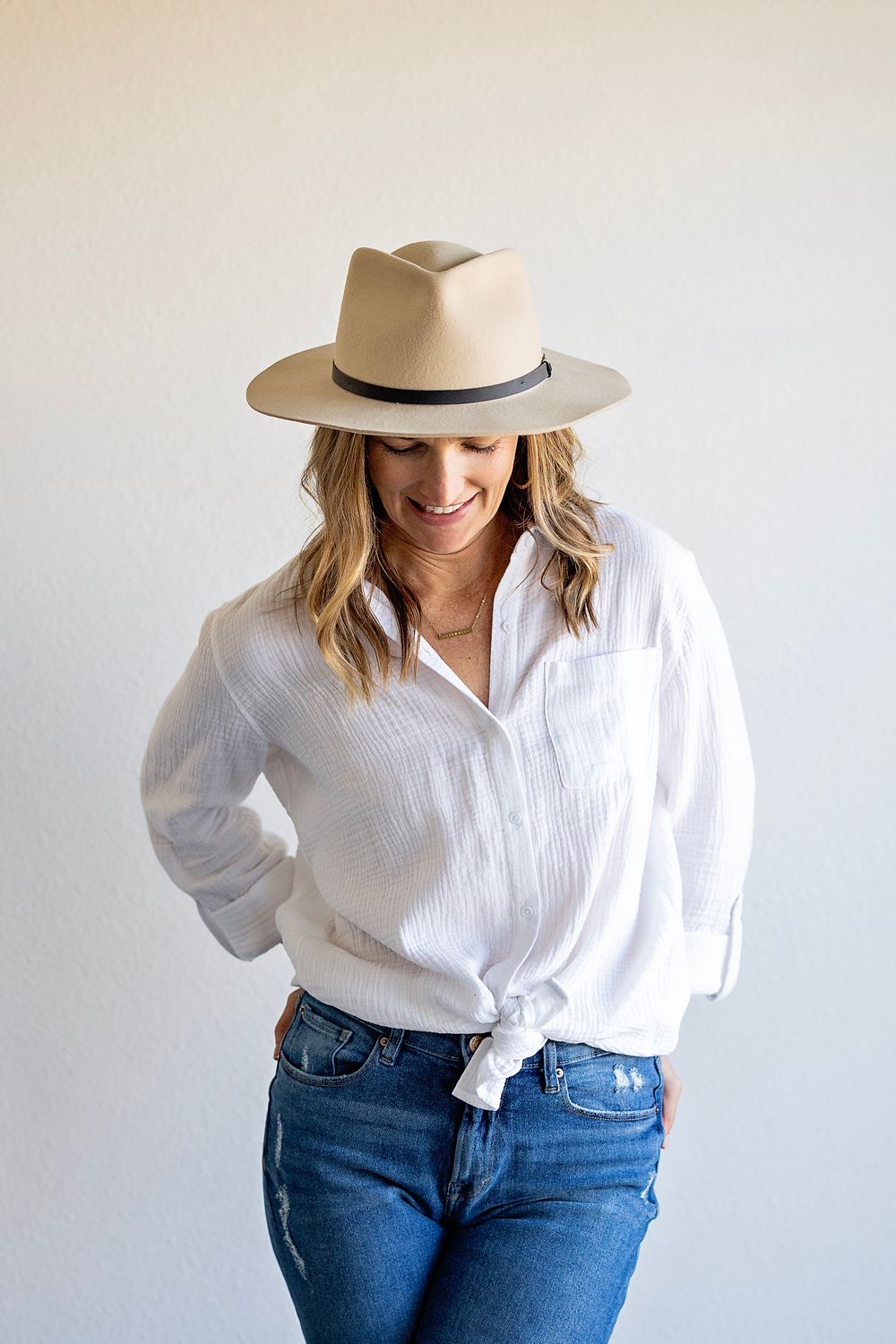A person wearing a beige fedora hat, white button-down shirt, and blue jeans poses against a light wall.