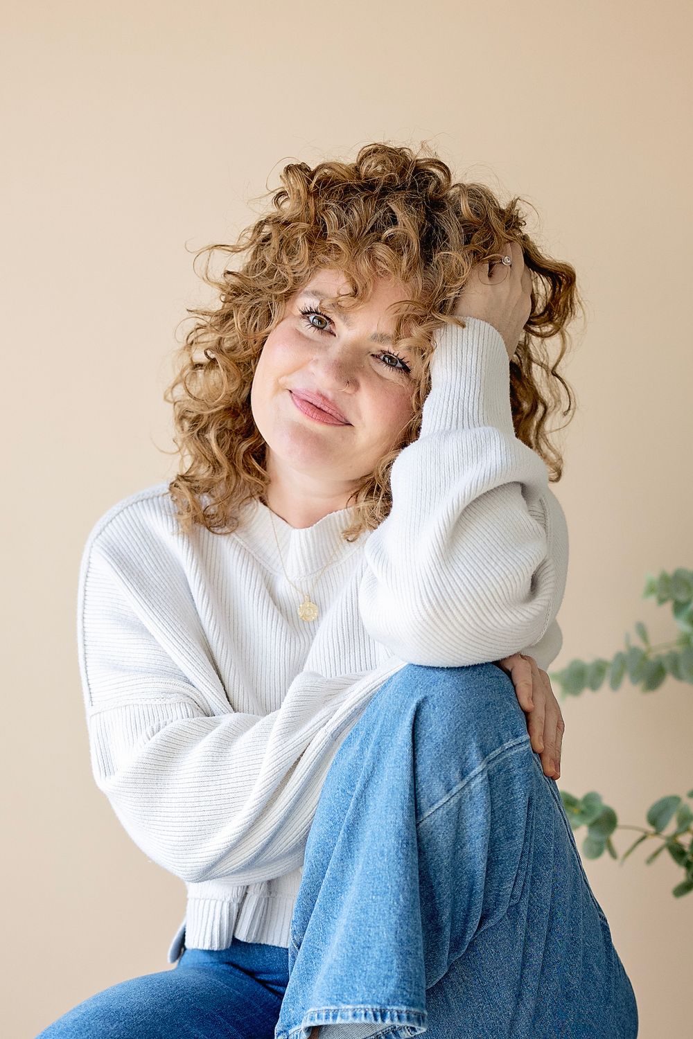 Person wearing white knit sweater and blue jeans poses against neutral wall with plants in background.