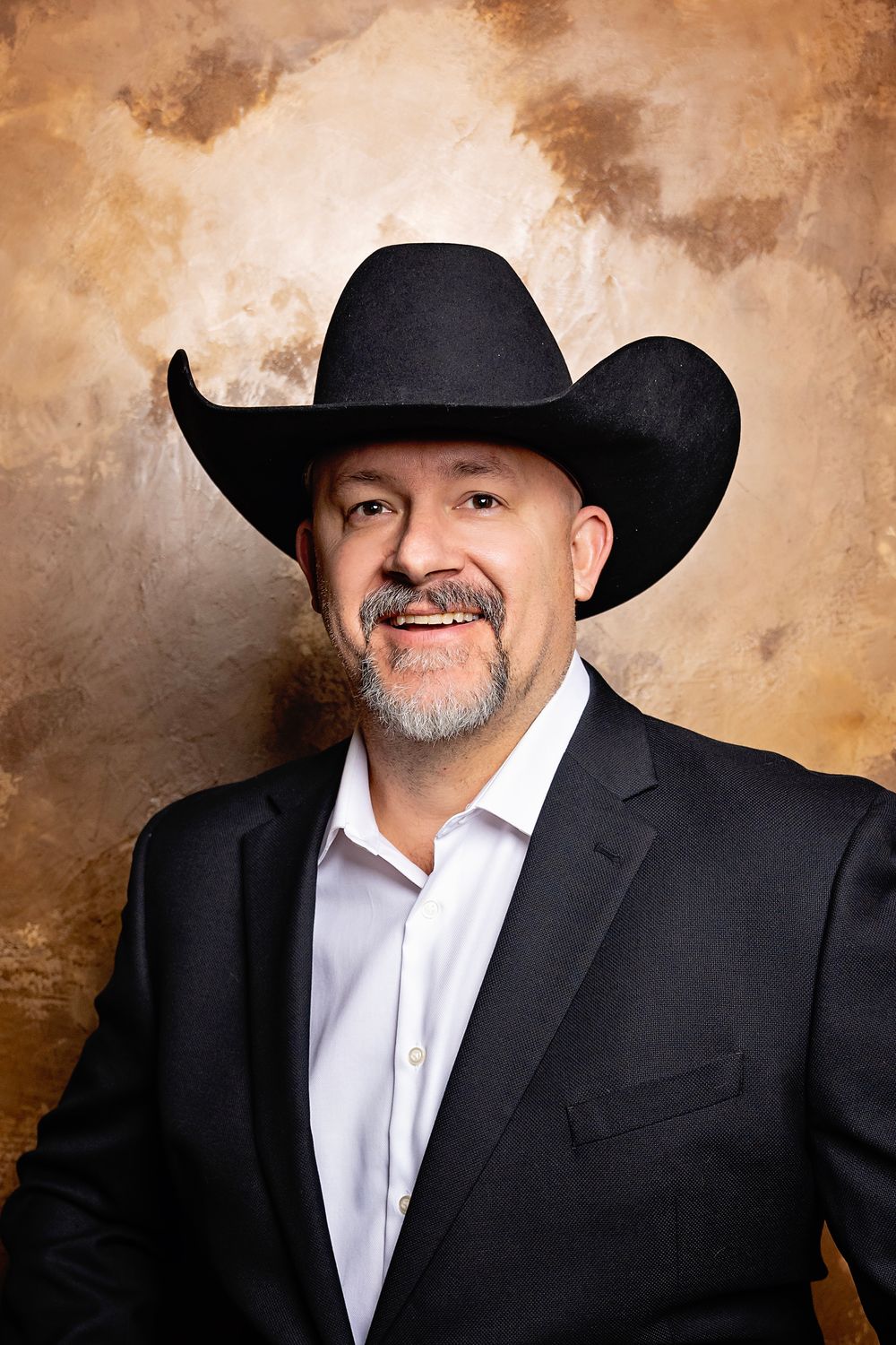 Professional headshot portrait with black cowboy hat and suit against textured brown background.