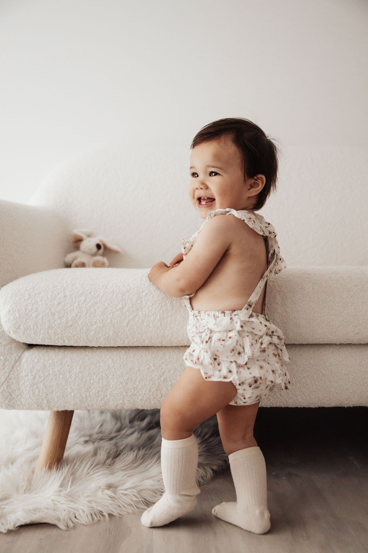 Happy baby in white outfit sitting by sheer curtains in bright room.