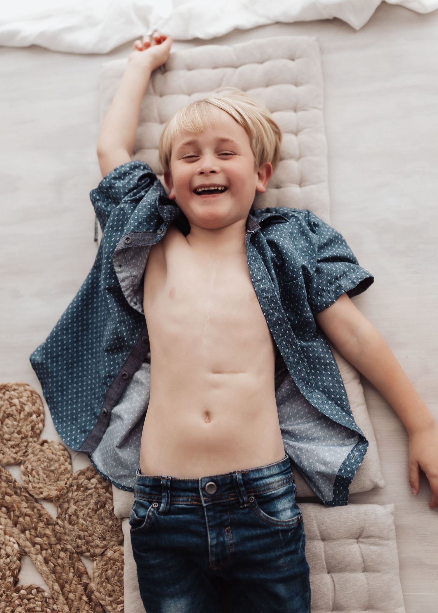 A young child lying on a quilted surface wearing an unbuttoned polka dot shirt and jeans while laughing joyfully.