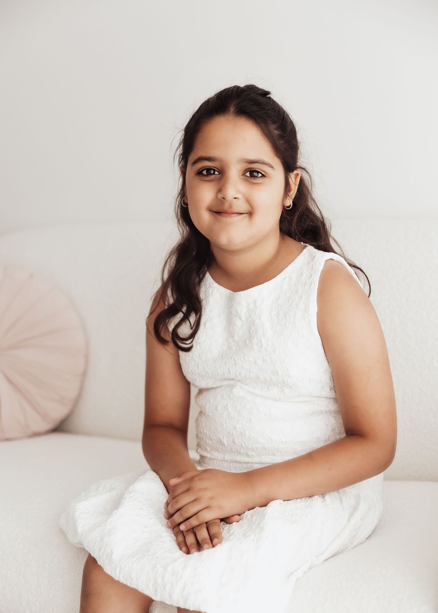A young child wearing a white sleeveless lace dress sits on a light colored couch with a pink pillow in the background.