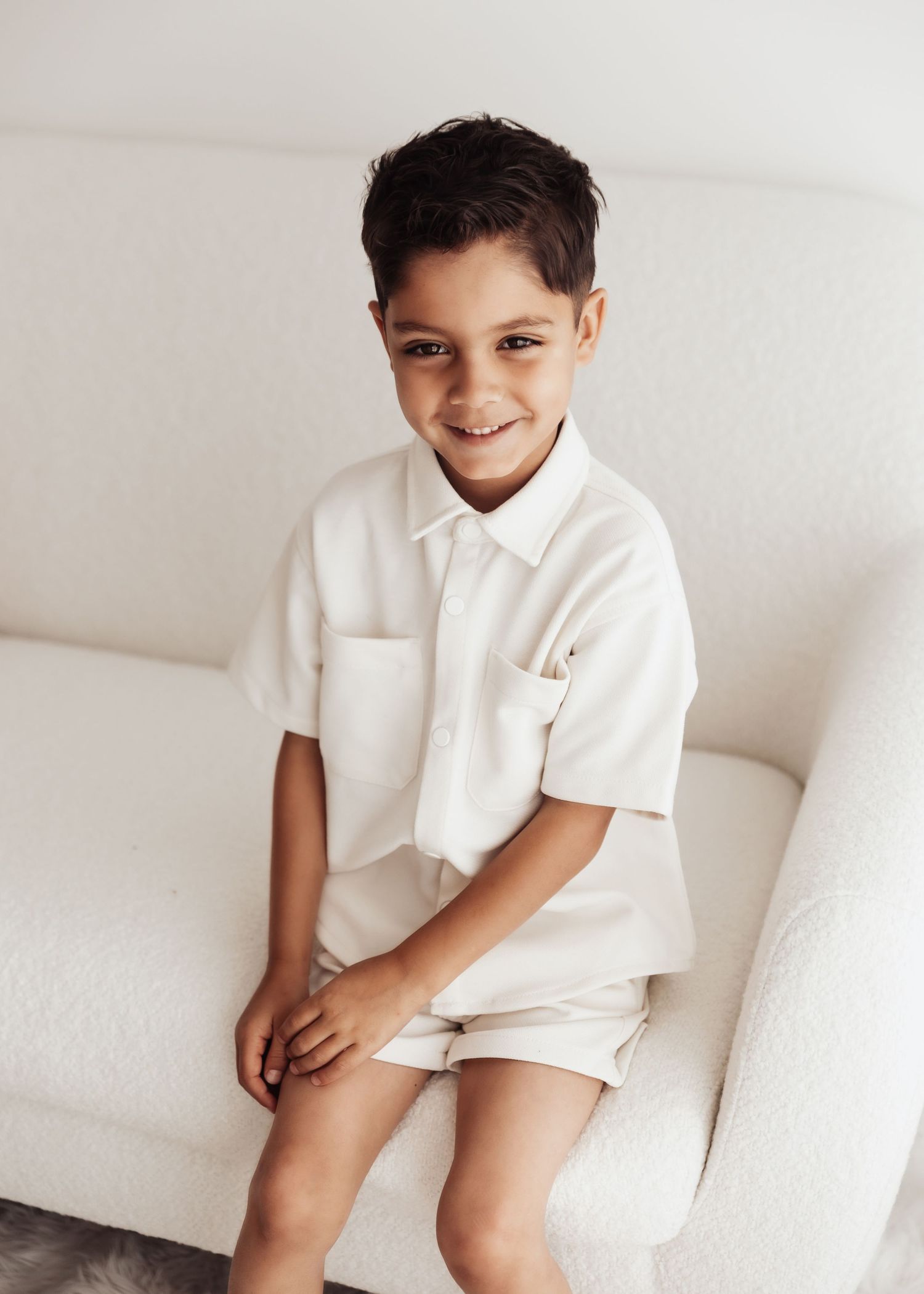 A child in a white polo shirt and shorts sits casually on a white couch against a light background.