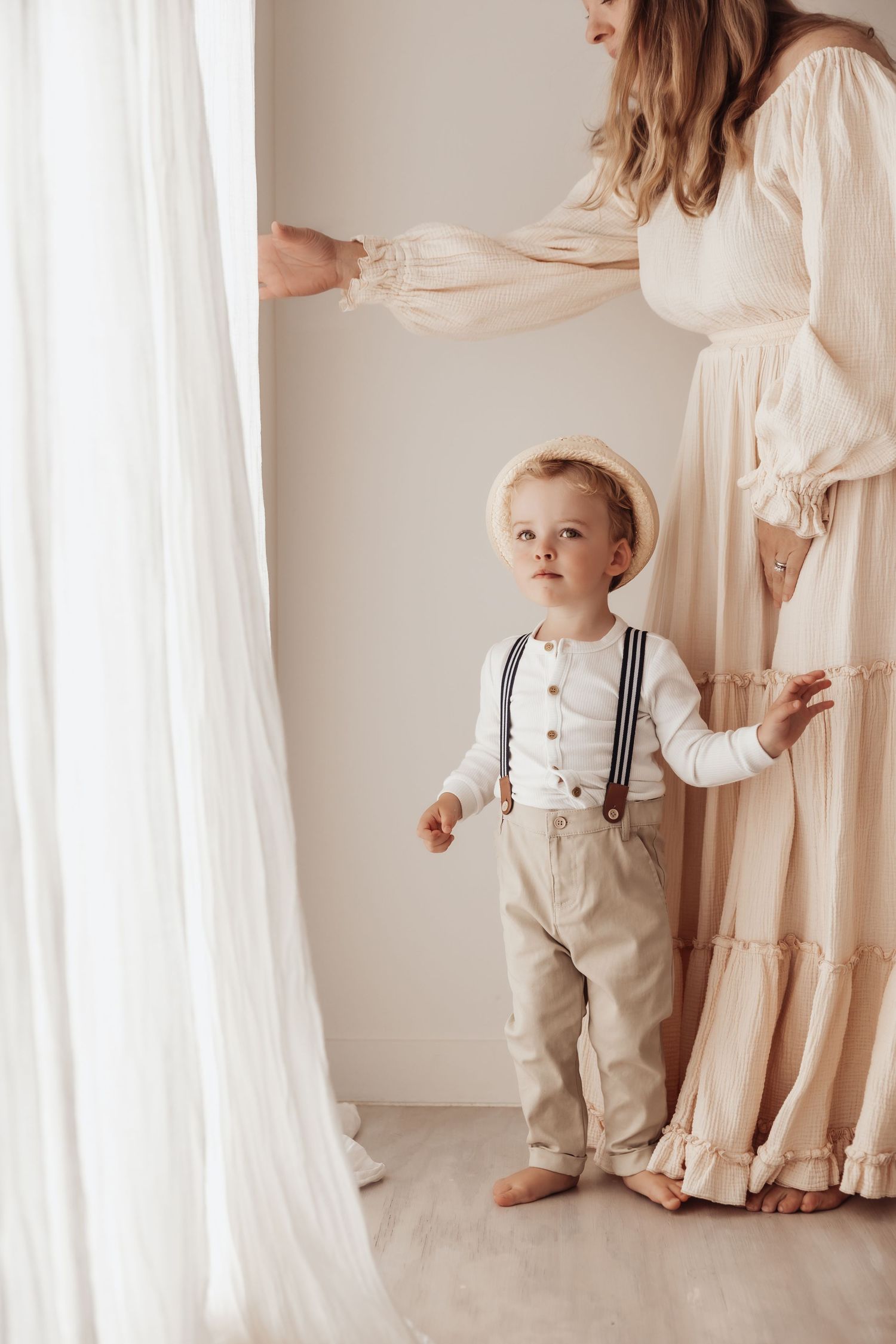 A small child in white suspenders and linen outfit stands beside flowing white curtains and soft beige fabric.
