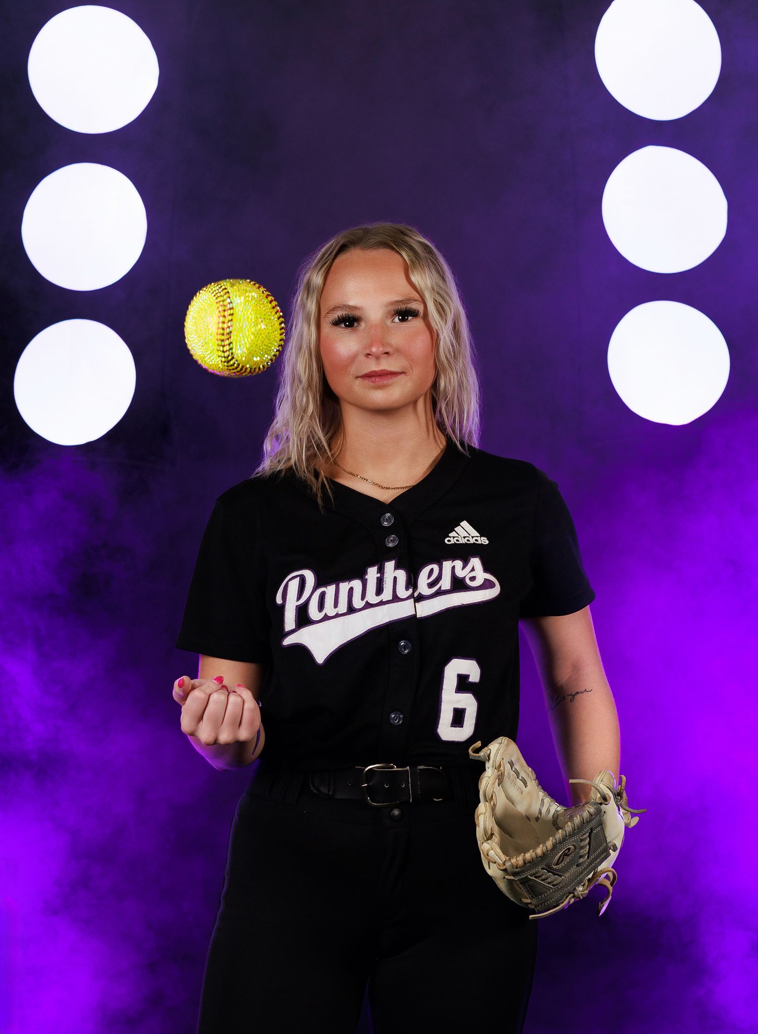 Softball player in black Panthers uniform number 6 poses with ball and glove against purple backdrop with spotlights.