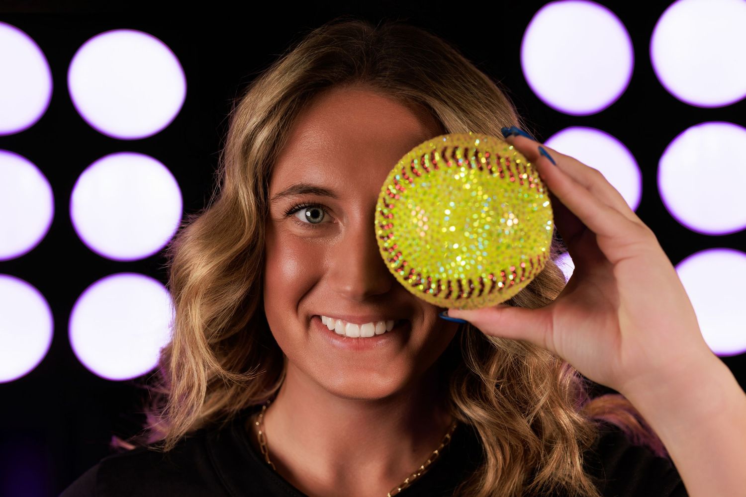 Softball player poses with yellow ball covering one eye against bright white circular lights in background.