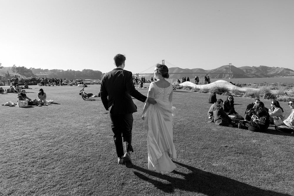 A couple in wedding attire walks together on a grassy hillside with scenic mountain views in the background.