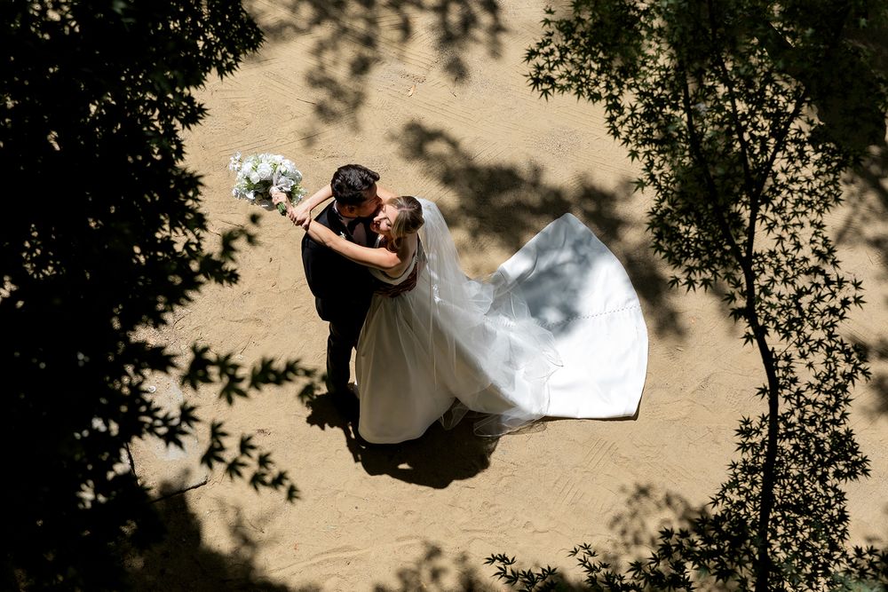 Aerial view of couple embracing during wedding ceremony with flowing white dress and natural tree framing.