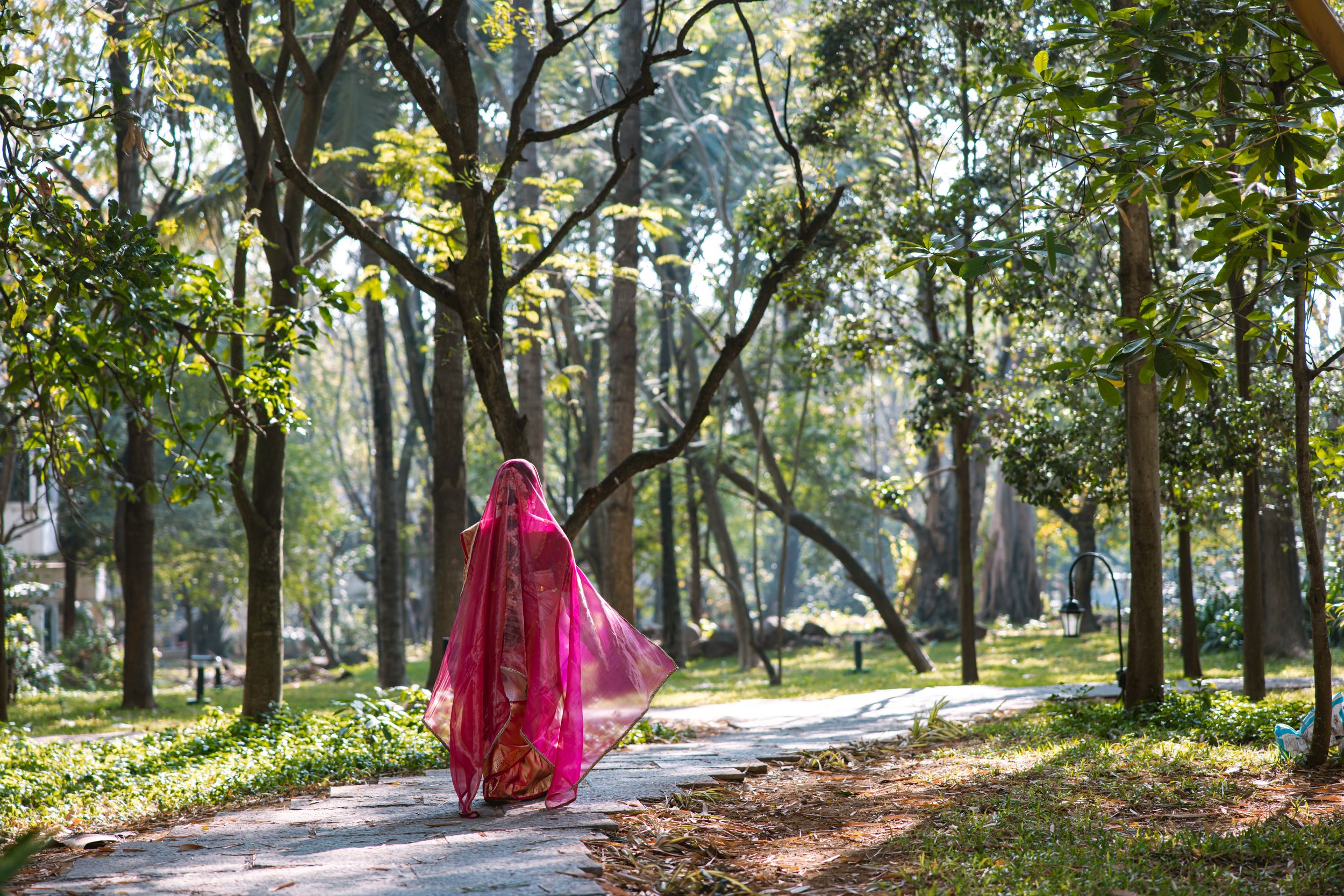 Woman in vibrant red dress walking through a sunlit wooded park.