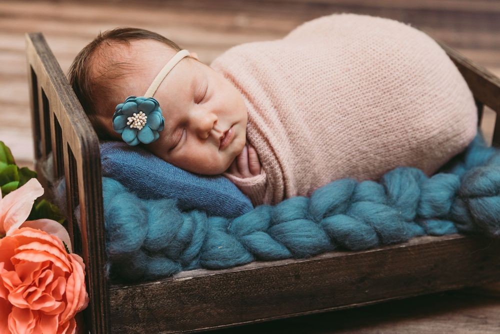 Sleeping newborn wrapped in pink blanket on blue knit backdrop with coral rose accent in wooden bed frame.