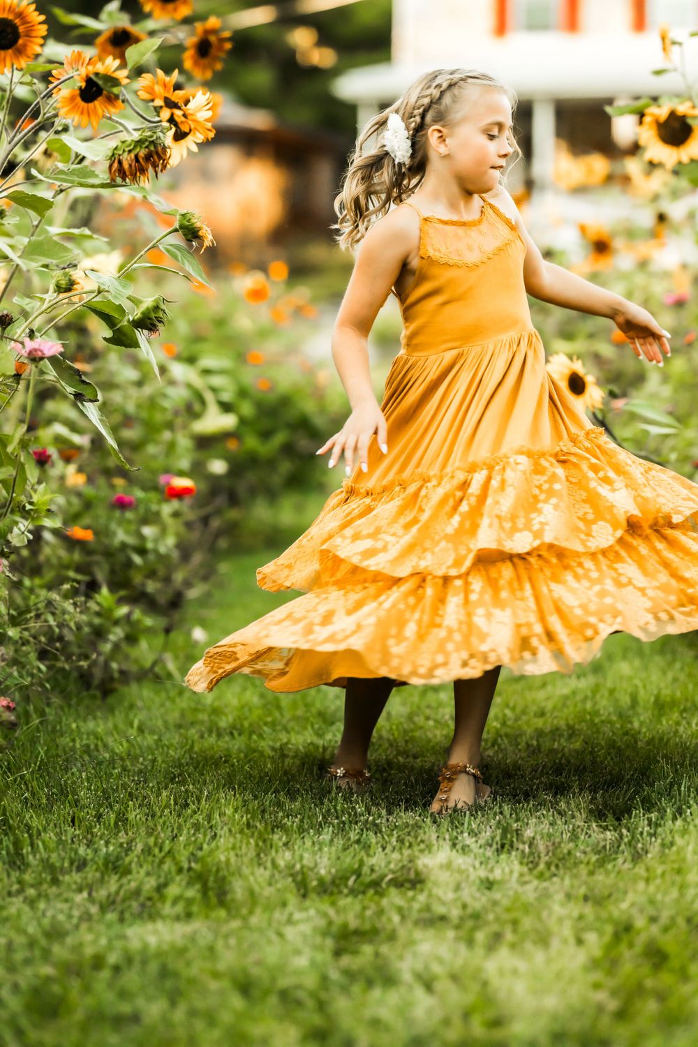 A child in a flowing yellow dress twirls among sunflowers in a garden setting.