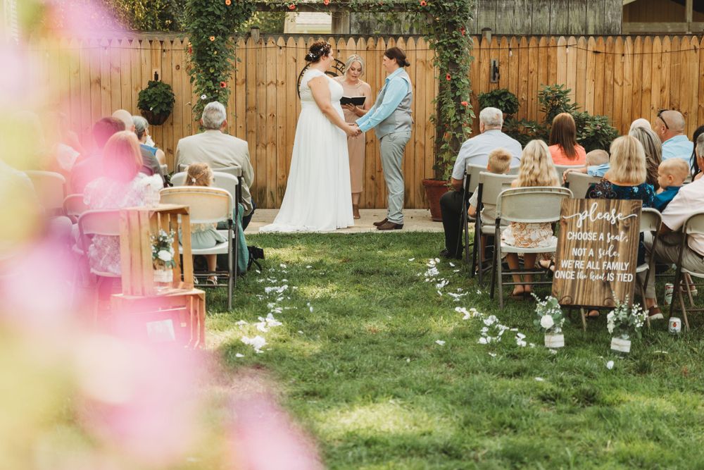 An intimate backyard wedding ceremony with guests seated on chairs and pink flowers in the foreground.