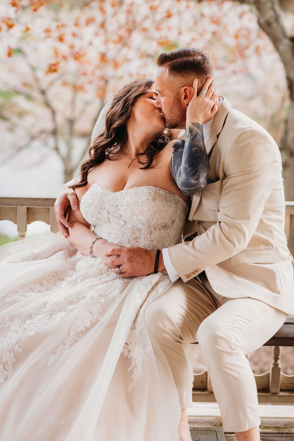 Romantic couple in wedding attire shares tender moments on bench surrounded by spring blossoms.