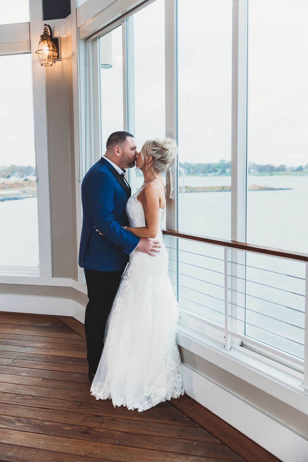 Newlyweds share a romantic moment by large windows overlooking the water.