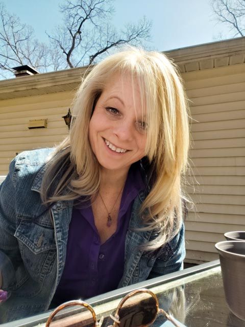 A person wearing a denim jacket and purple shirt sits outdoors on a sunny day with sunglasses resting nearby.
