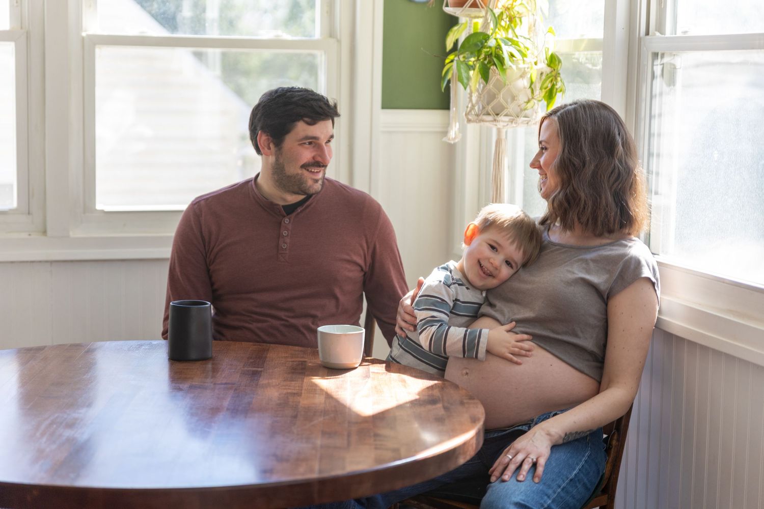 Maternity - Expecting family smiling together at a wooden kitchen table in a bright sunlit home.