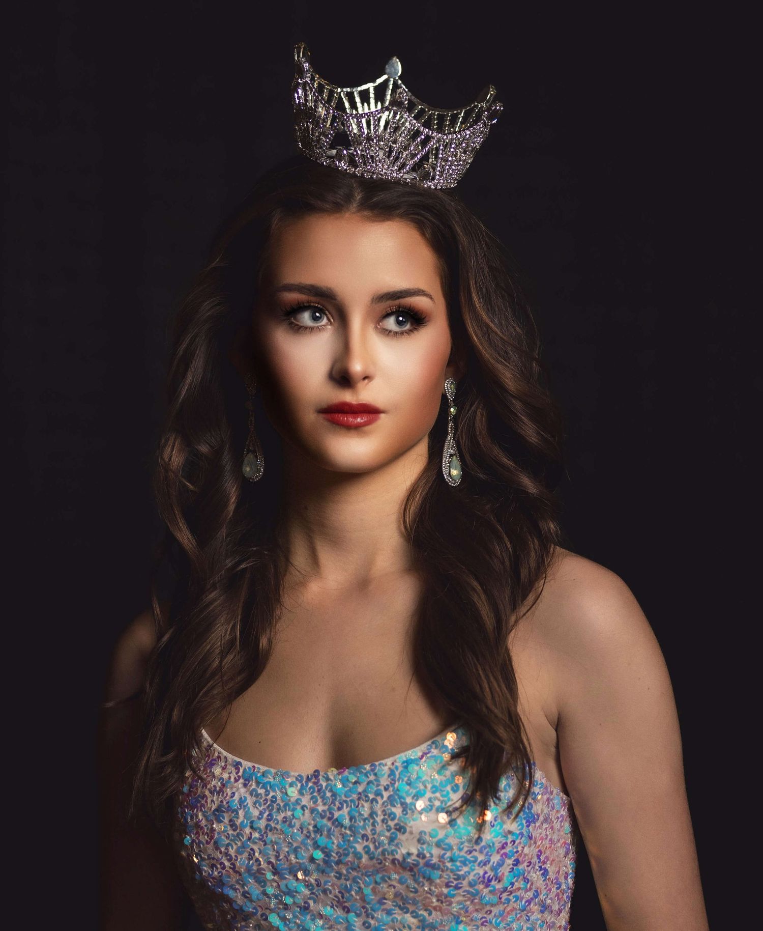 Dark elegant portrait showing a sparkling crown and turquoise beaded gown against a black background.