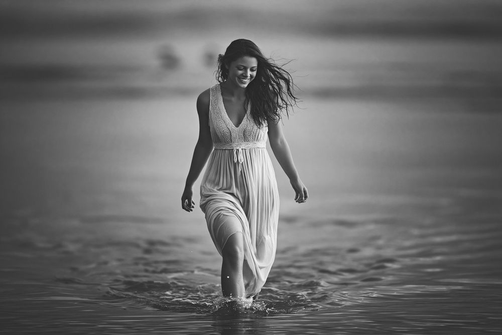 Black and white photo of a smiling woman in a white dress walking through shallow ocean waves on a beach.