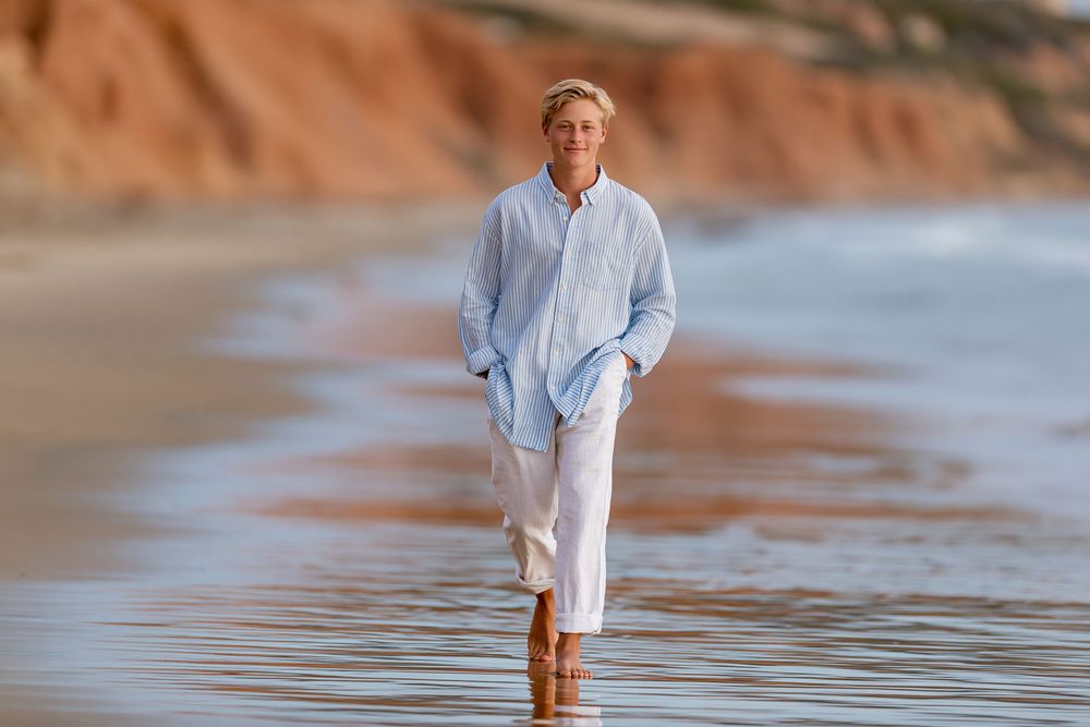 Man in light blue linen shirt and white pants walking barefoot on a sandy beach with red cliffs.