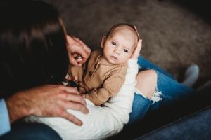 A small infant in a tan outfit looks up while being held on someone's lap.
