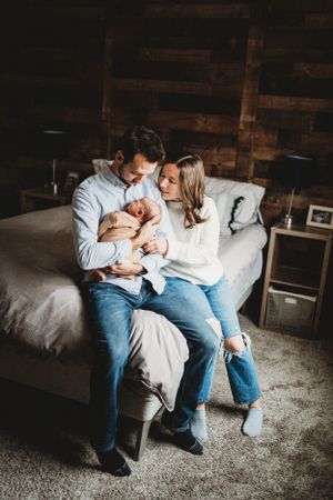 A couple sits on their bed holding their newborn baby in a rustic bedroom setting.