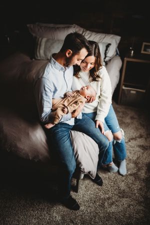 A couple sits together on a grey couch while holding their baby in a tan outfit.