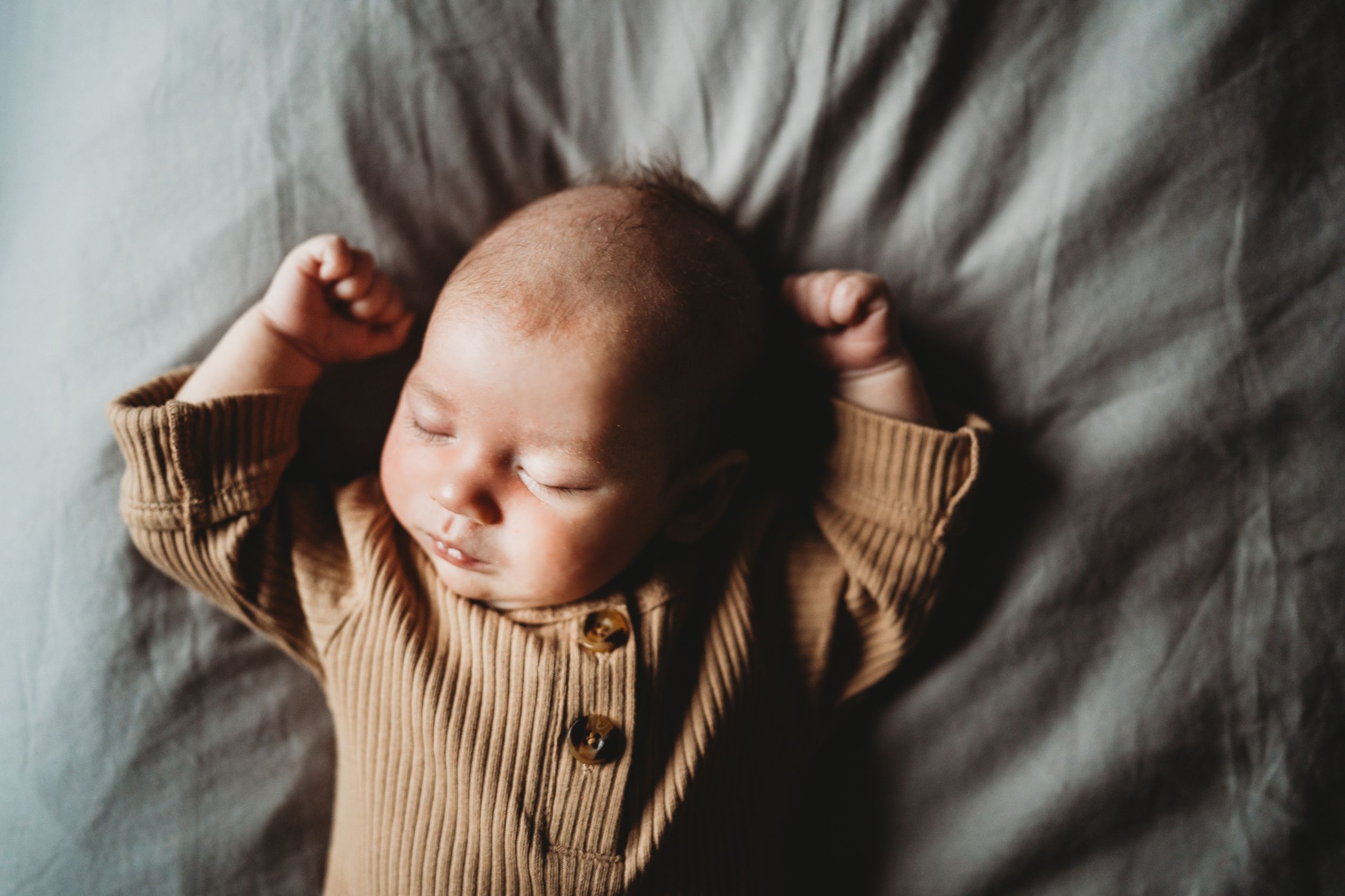 Sleeping infant wearing beige knit outfit stretches arms overhead on bed.