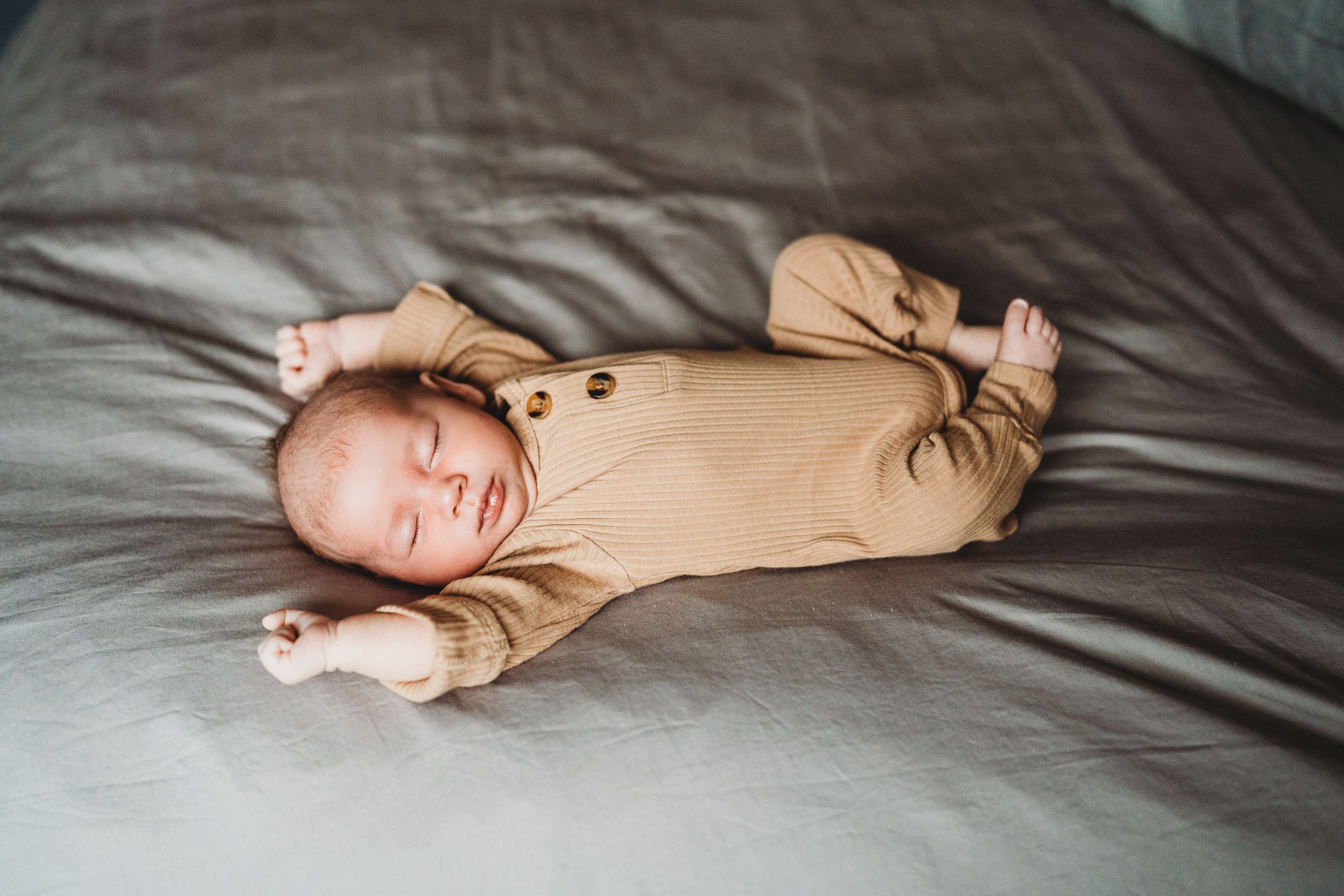 Newborn baby in tan onesie stretches while lying on gray bedding.